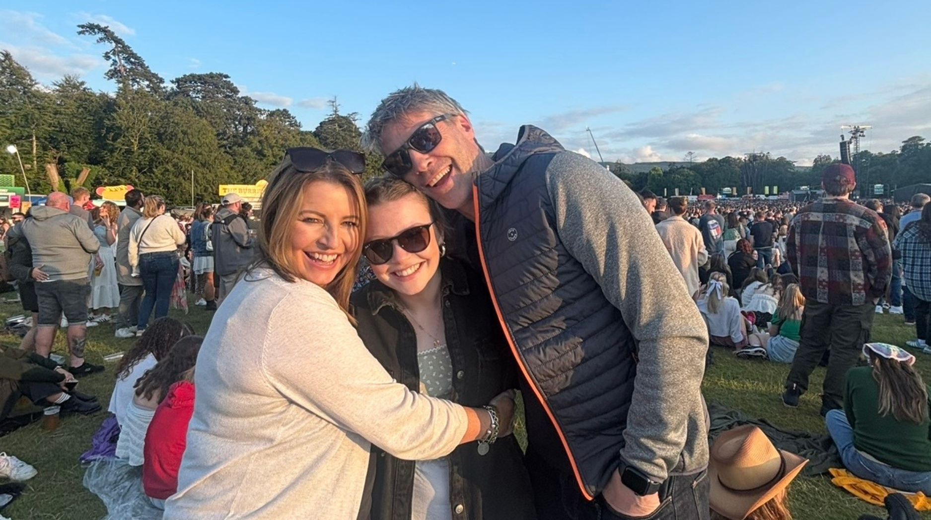 Ella and her parents smiling at an outdoor concert on a sunny day with a blue sky