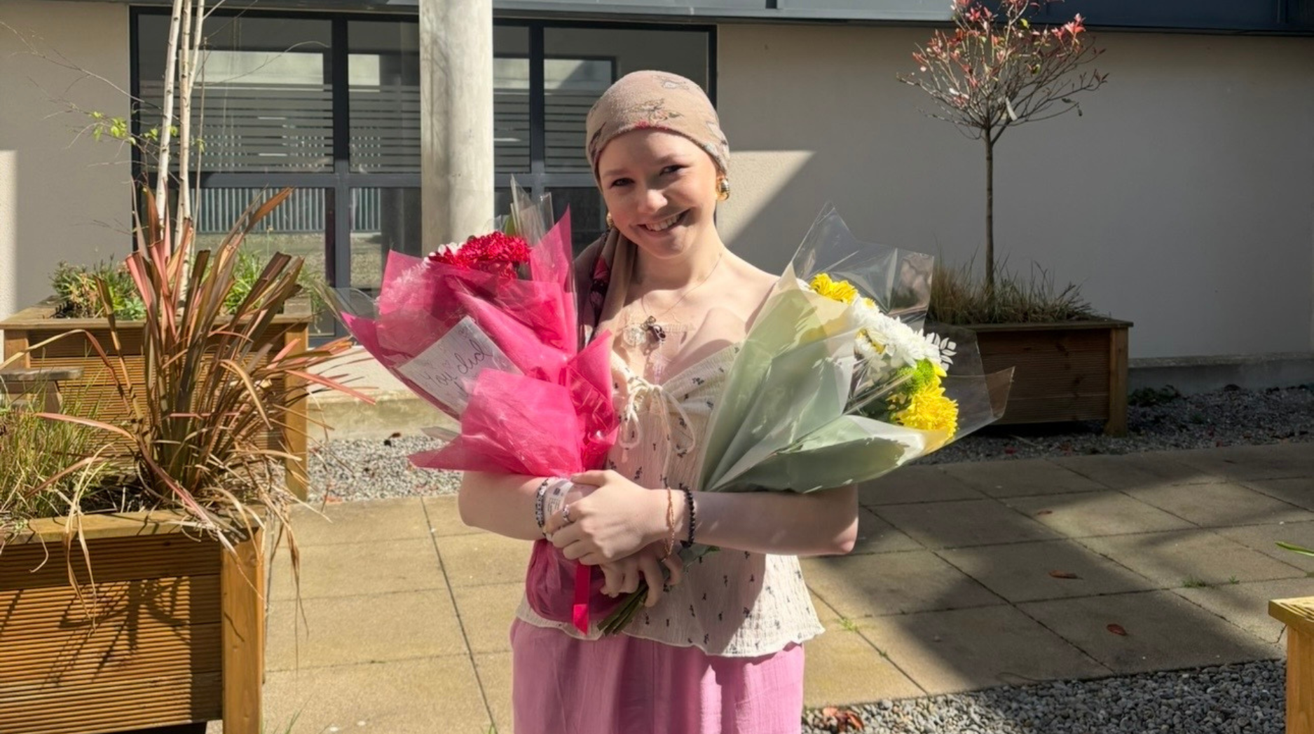Ella standing outside in the sun holding two colourful bunches of flowers, one in each hand