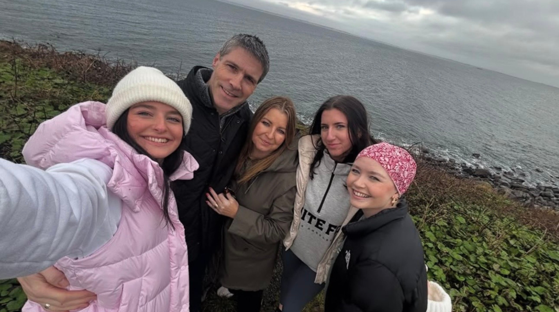 Ella with her parents and two sisters, standing close together and smiling with a sea view behind them