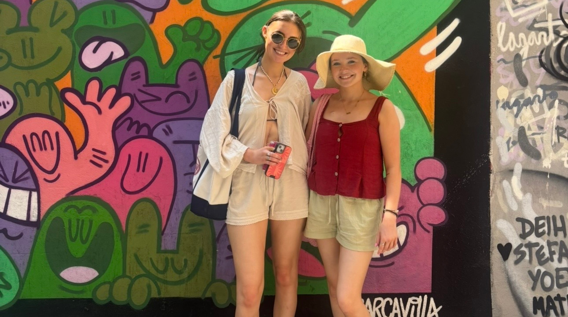 Ella and a friend smiling in front of a vibrant, colourful mural on holiday with the sun shining.