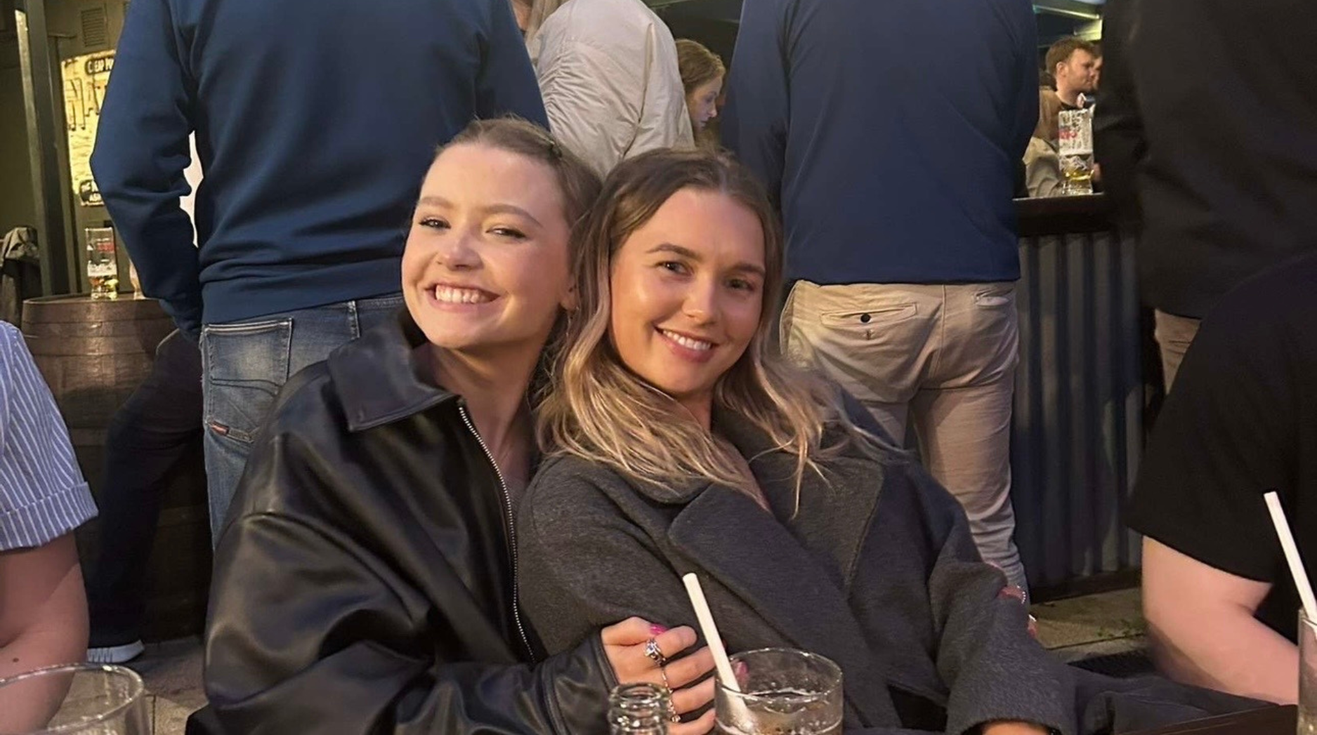 Ella and a friend sitting close together with her hand resting on her friend's arm, both smiling.