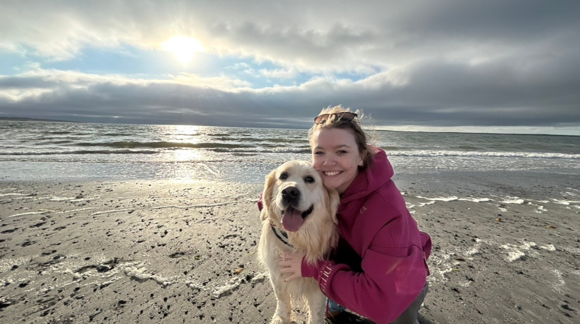 Ella and her dog at the beach, smiling while kneeling on the sand with the sea in the background