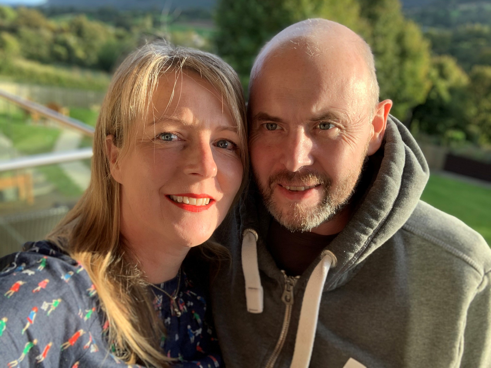 The image shows Lisa and John, husband and wife, smiling at the camera with trees in the background.