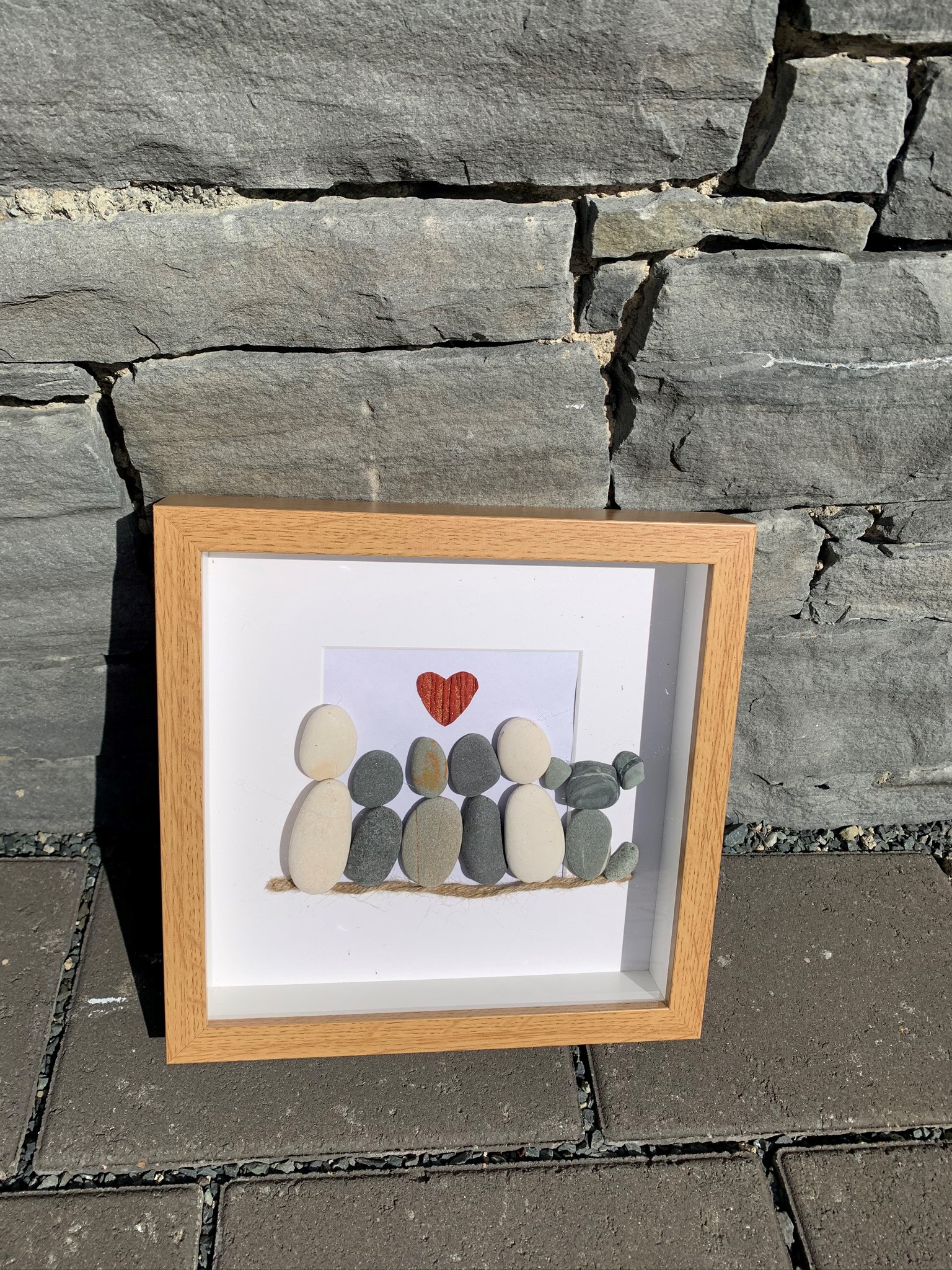 The image shows an art piece in a wooden frame. A family of 5 are depicted, made up of different coloured stones with a red heart above their heads.
